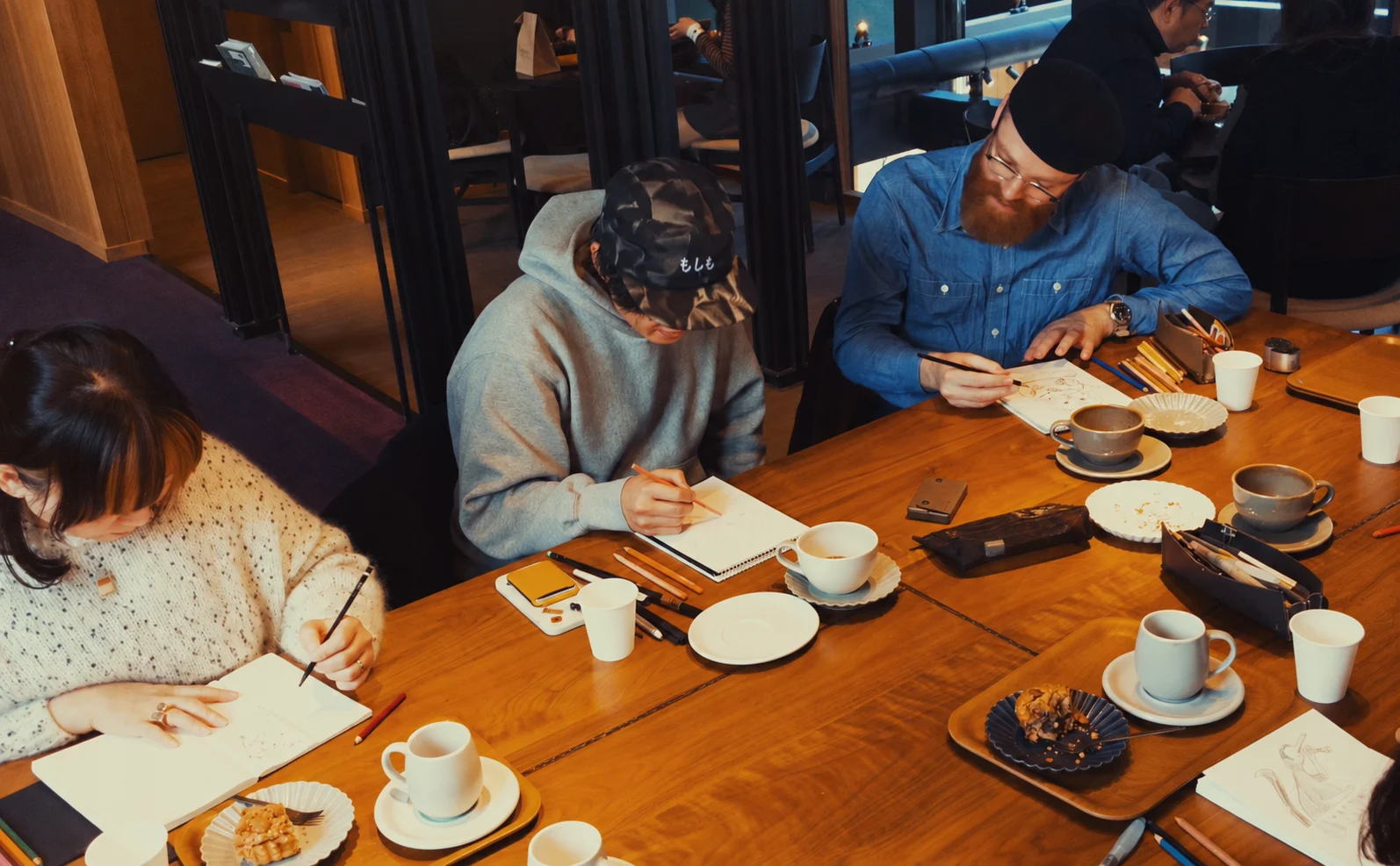 People drawing in sketchbooks around a cafe table with coffee and pencils.